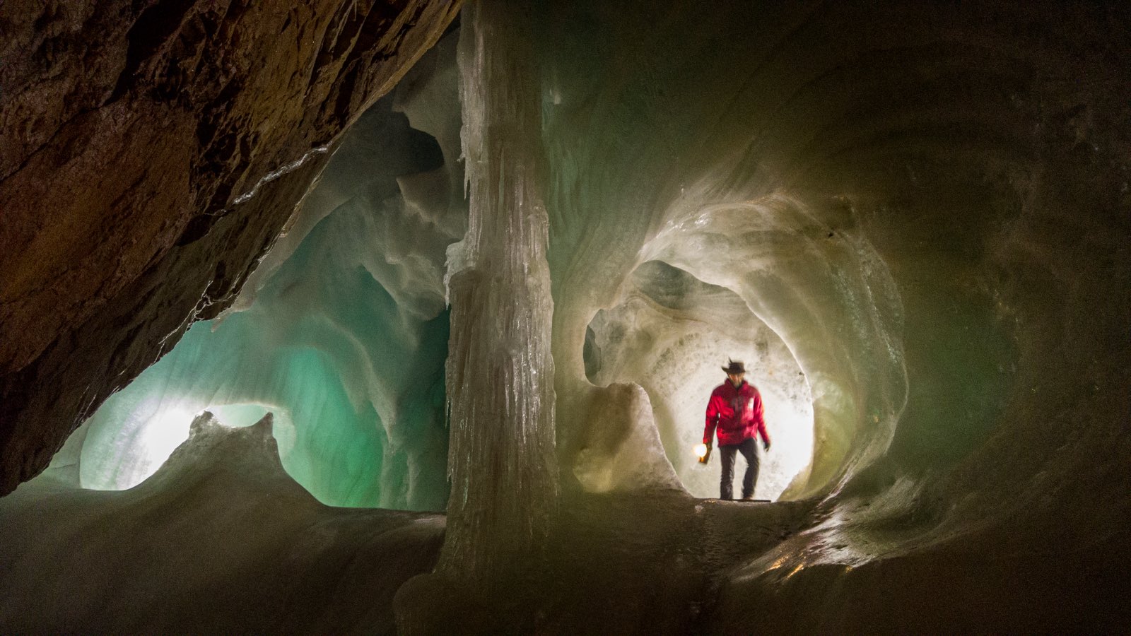 A lone figure in a red jacket, holding a lantern, standing deep inside a glowing blue-white ice cave