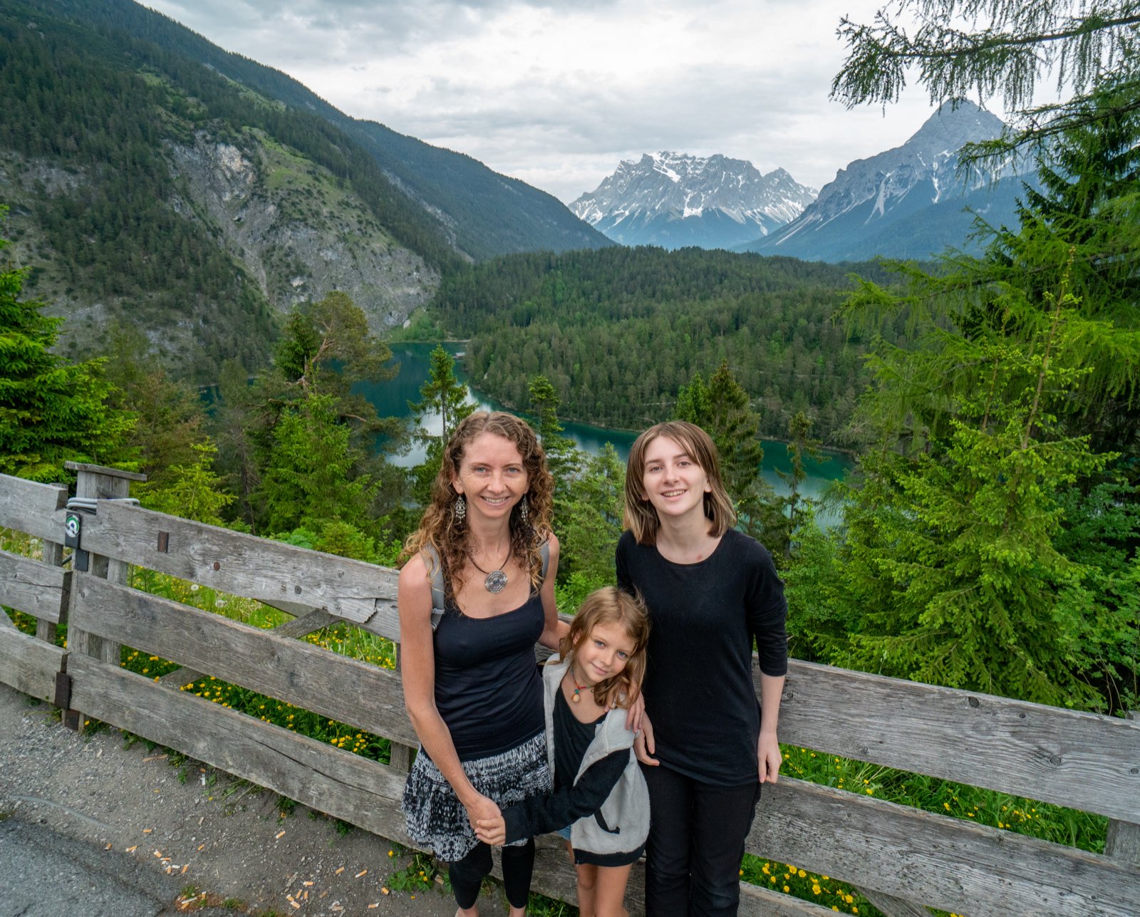 Jennifer and the two younger girls smiling by a wooden fence above a turquoise lake ringed with forest and peaks
