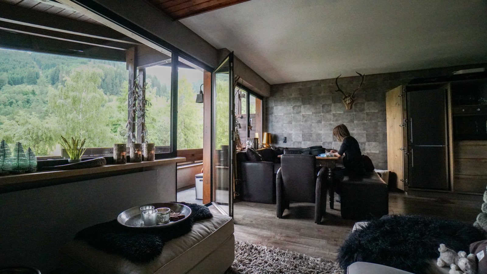 A girl writing at a table in a dim, wood-and-stone alpine apartment, with tall windows opening onto a green hillside