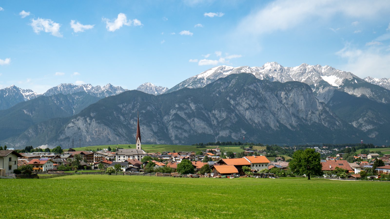 A Tirolean village with a church steeple set against snow-streaked Alps under a blue sky