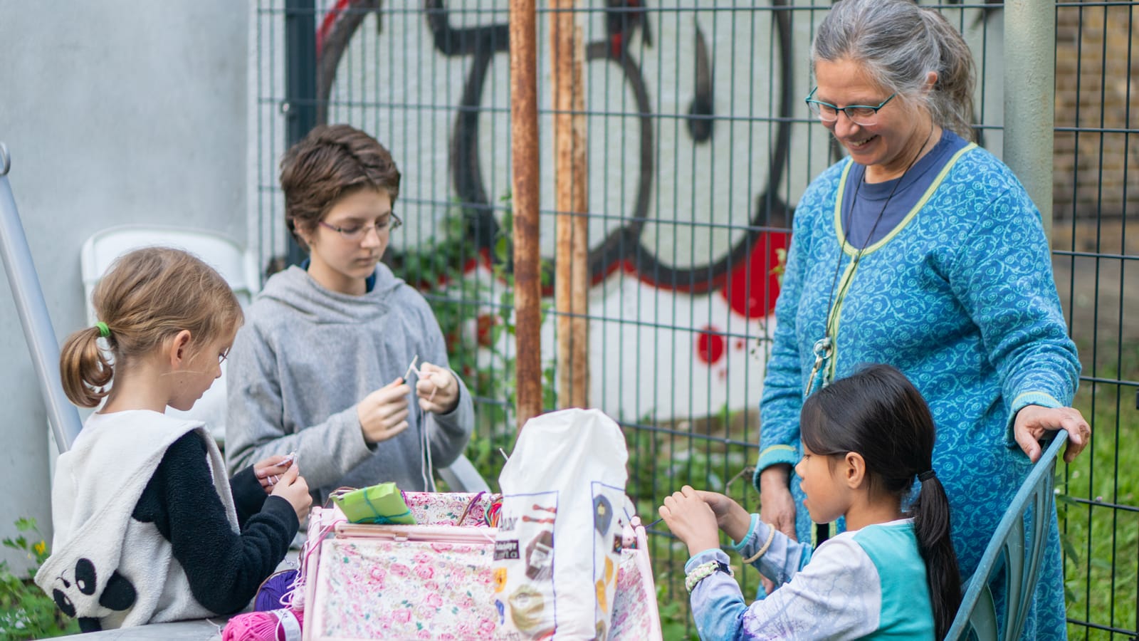 Kids gathered around an older woman, learning a handwork project outdoors