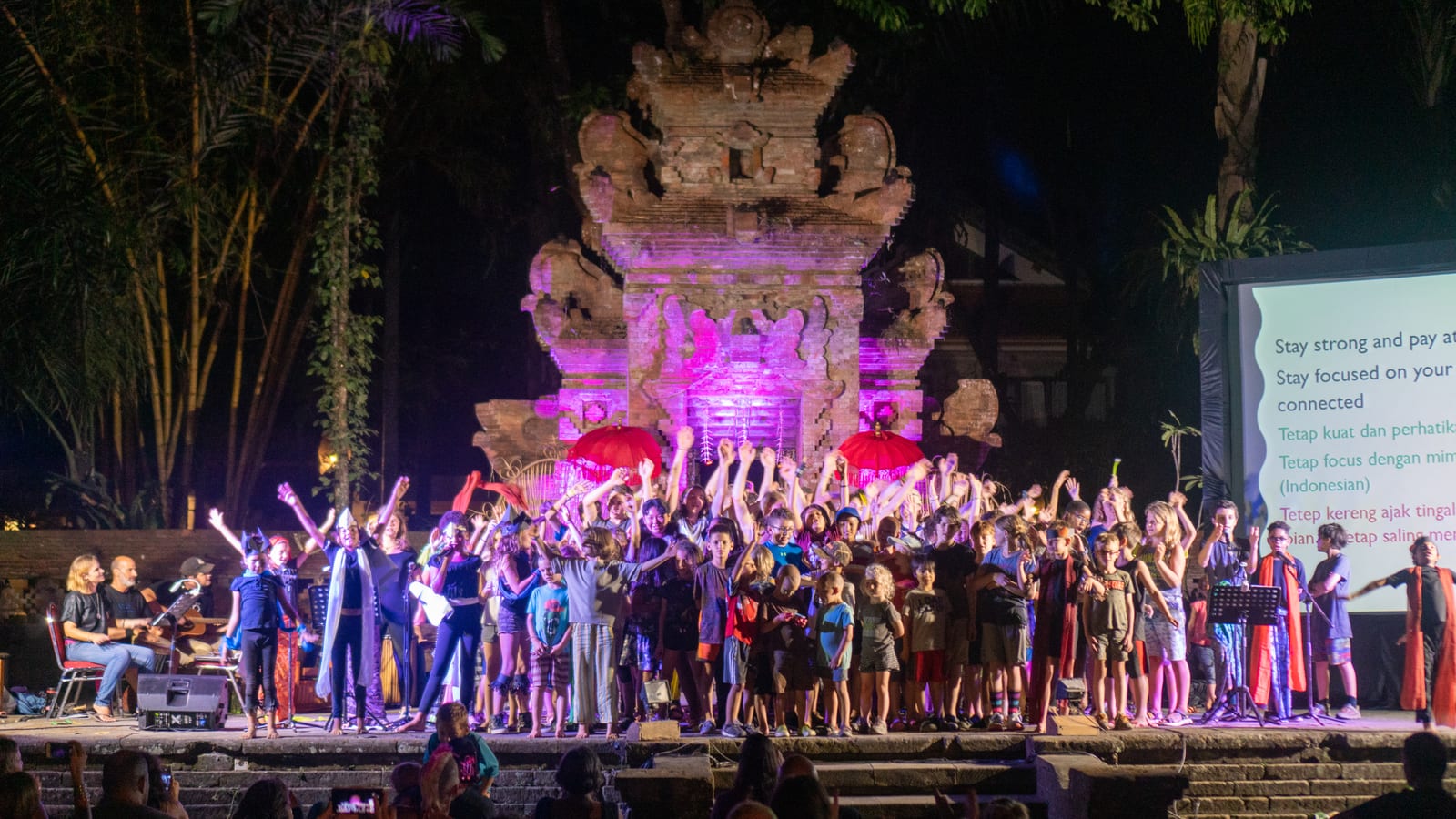 Our whole community gathered on stage, singing together in front of a purple-lit Balinese temple gate on the closing night