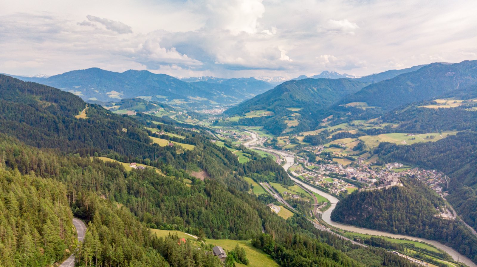 A high view down a broad green Alpine valley, a river winding past a town with a castle on a wooded bluff