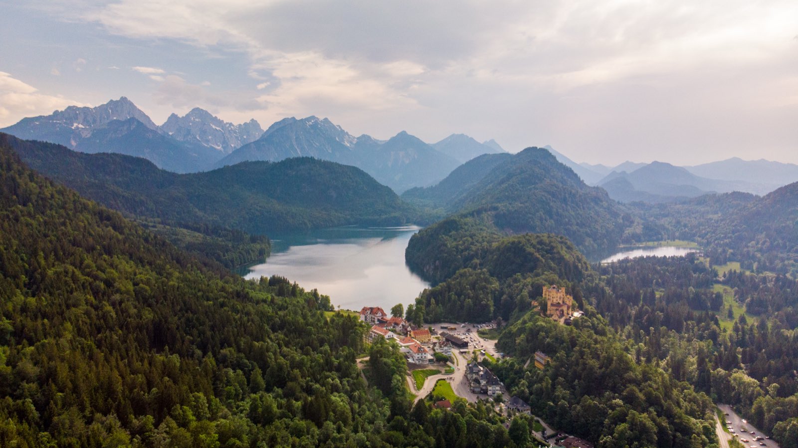 An aerial view of the yellow Hohenschwangau castle beside the Alpsee lake, ringed by forested Alps